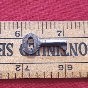 May include: A small, antique metal key with an oval head and a ridged shaft, resting on a wooden ruler. The ruler shows inch measurements. The key is likely for a small lock or piece of furniture.