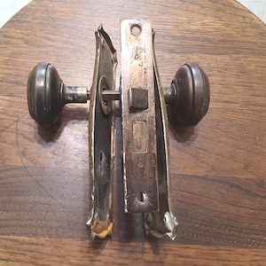 May include: An antique door knob set with a tarnished bronze finish. The set includes two round knobs and a mortise lock with a rectangular faceplate. The hardware shows signs of age and wear, with a dark patina.
