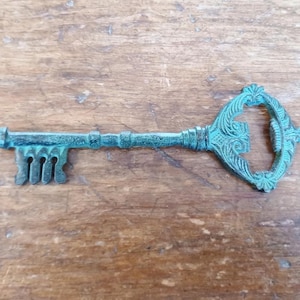 May include: A teal-coloured, vintage-style key with an aged patina. The key has an ornate, decorative head with scrollwork and a detailed bit. The metal key is on a wooden surface.