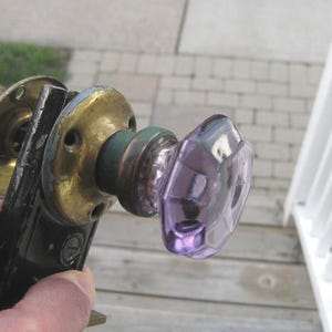 May include: Vintage door handle with a faceted, purple glass knob. The knob is set on a brass plate with a black metal backplate. The hardware shows signs of age and wear, with a green collar.