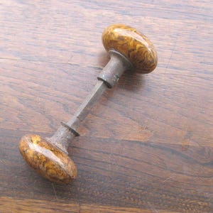 May include: Vintage door handle featuring oval knobs with a marbled effect in brown and yellow tones. The handle has a metal construction with a rectangular stem. The hardware shows signs of age, indicating it is an antique piece.
