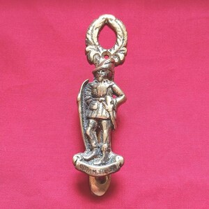 May include: A brass door knocker featuring a figure dressed in historical attire, including a hat and tunic, standing on a base with the engraved text Robin Hood. The top of the knocker is shaped like a decorative wreath with a central opening.