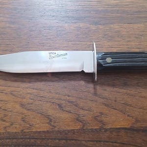 May include: A fixed-blade knife with a silver-toned steel blade and a black handle. The blade features the engraved text Utica Sportsman U.S.A. The handle is secured with two brass-coloured rivets and includes a metal guard between the blade and handle.