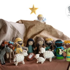 Pode incluir: Um presépio de crochê artesanal com figuras da Sagrada Família, reis magos, anjos e animais. As figuras são de várias cores, incluindo bege, azul e verde, com uma estrela amarela acima. A cena é ambientada em um fundo marrom.