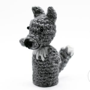 May include: A grey crocheted wolf puppet with black eyes and nose, and white accents on the chest and paw. The handmade toy is set against a white background. The wolf has pointed ears and is facing to the left.
