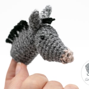 May include: A handmade crochet donkey head in shades of gray, with black accents for the mane and ears. The donkey has a light pink nose and is held against a white background. The product is a craft item.