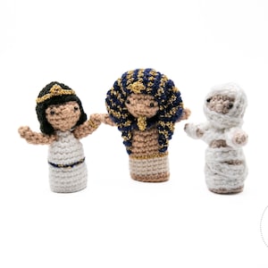 Pode incluir: Três fantoches de dedo feitos à mão em crochê: uma rainha egípcia, um faraó e uma múmia. A rainha usa um vestido branco e detalhes dourados. O faraó tem um cocar azul e dourado. A múmia é embrulhada em branco. Tudo em um fundo branco.