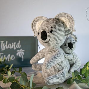 Puede incluir: Dos koalas de ganchillo, uno grande y otro pequeño, en tonos grises. El koala más grande tiene su brazo alrededor del más pequeño. El fondo incluye un letrero de madera que dice "Island Time" y una ramita de hojas verdes.