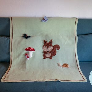 以下が含まれることがあります： 茶色の縁取りが施された、刺繍された森の生き物が特徴の、薄緑色のニットブランケット。 ブランケットには、リス、キノコ、カタツムリ、蝶がいます。 ブランケットは、枕付きのティール色のソファに飾られています。