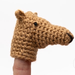Op de afbeelding: Een gehaakte vingerpop in de vorm van een kameelkop. De pop is gemaakt van bruin garen met een zwart oog en een donkerbruine snuit. De kameelpop zit op een vinger tegen een witte achtergrond.
