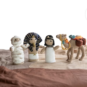May include: Crocheted figures of an Egyptian pharaoh, a mummy, a woman, and a camel with saddlebags. The figures are beige, white, and brown, with blue, gold, and black accents. The figures are arranged on a brown and beige fabric background.
