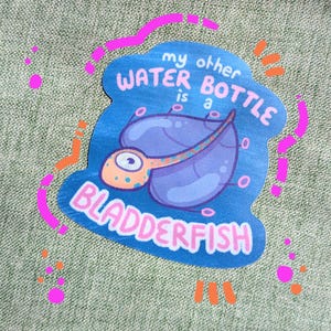 Può includere: Un adesivo blu con la scritta "my other WATER BOTTLE is a BLADDERFISH". L'adesivo presenta un'illustrazione a cartoni animati di un pesce vescica in viola e arancione, con accenti rosa e arancioni.