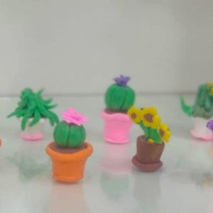 Puede incluir: Una colección de coloridas plantas en macetas de arcilla en miniatura. El surtido incluye cactus, girasoles y otros arreglos florales en macetas rosas, naranjas y marrones. Las plantas están dispuestas sobre una superficie blanca.