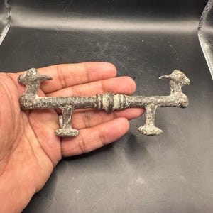 May include: A weathered, antique bronze door handle. The handle features a central bar with decorative ridges and bird-shaped finials at each end. The handle is supported by two rectangular posts. The overall design suggests a vintage or historical aesthetic.