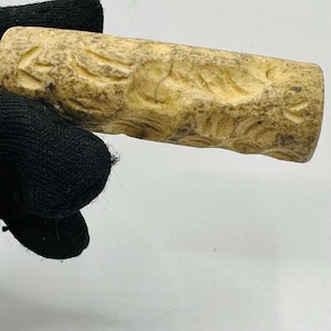 May include: A carved, cylindrical object with a light tan color and intricate floral patterns. The object is held against a white background, with a black glove partially visible.