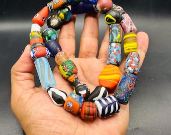 May include: A strand of multicolored glass beads featuring various shapes, sizes, and patterns. The beads display a wide array of colors including red, blue, yellow, green, black, and white, with some showing striped, speckled, or floral-like designs.