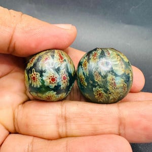 May include: Two spherical glass beads featuring a dark green base colour with an intricate millefiori pattern. The surface displays numerous small, repeating floral-like motifs in shades of white, light blue, and red, creating a textured appearance.