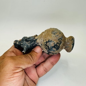 以下が含まれることがあります： 片方の端に暗い動物の頭が付いた、小さく、アンティーク風の陶器の容器。容器の本体はまだら模様の茶色と黒色です。このオブジェクトは手で持たれています。