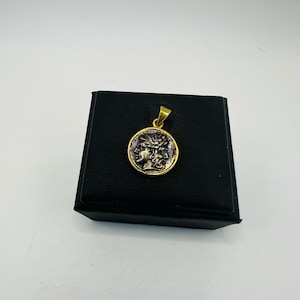 May include: A gold pendant with an antique coin design. The coin has a detailed relief of a face and other figures. The pendant is displayed on a black box against a white background.