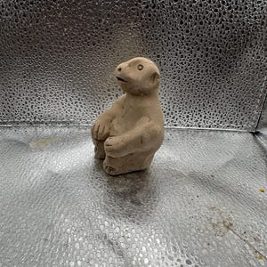 May include: A small, tan-colored ceramic sculpture of a seated animal with a rounded body, short legs, and a small head. The sculpture has a smooth texture and is set against a textured silver background.