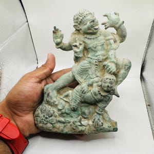 May include: A weathered, green-toned statue depicting a seated figure atop another smaller figure. The larger figure has a detailed face and is adorned with jewellery. The statue appears to be made of a metal or stone material.