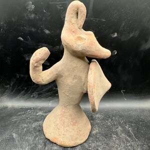 Pode incluir: Estatueta de terracota de uma figura animal estilizada com um focinho longo, orelhas grandes e um objeto em forma de escudo. A figura está em pé sobre uma base alargada, com um braço levantado. A cor é castanho claro.