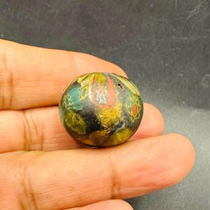 May include: A spherical bead featuring a dark base colour with intricate, multi-coloured floral and abstract patterns in shades of yellow, green, red, and blue. The surface of the bead displays a smooth, polished texture with a small central hole.