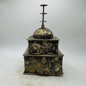 May include: A silver-toned, two-tiered box with intricate detailing. The box has embossed figures and floral patterns. A domed lid and a vertical, tiered finial crown the box, creating a unique decorative piece.