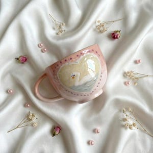 Può includere: Una tazza in ceramica rosa con un disegno a forma di cuore con un cigno. La tazza ha un manico curvo ed è decorata con piccole stelle viola. Lo sfondo include tessuto bianco, perle rosa e fiori secchi.