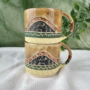 Pode incluir: Duas canecas de cerâmica empilhadas. Elas têm um esmalte marrom e bege com um desenho de montanha e árvore em preto, verde e branco. As alças são marrons e verdes. Adequado para bebidas quentes.