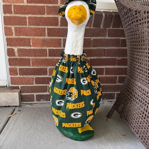 May include: A white goose statue wearing a dark green fabric outfit featuring the Green Bay Packers logo and text in yellow and white. The outfit includes a gathered dress covering the body and a matching ruffled bonnet on the head.