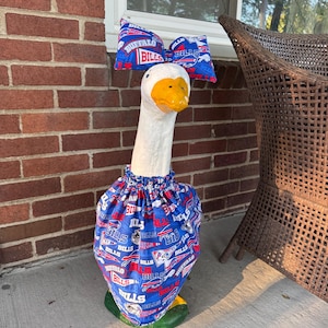 May include: A white goose statue wearing a blue, red, and white fabric outfit featuring the Buffalo Bills logo and pennant designs. The outfit includes a gathered dress around the body and a matching large bow attached to the top of the head.