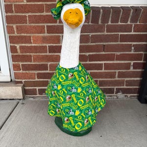 May include: A white goose statue with a yellow beak and a green and yellow patterned outfit. The outfit includes a matching bonnet and dress, both featuring the text "Oregon Ducks" and football graphics. The goose is standing on a green base.