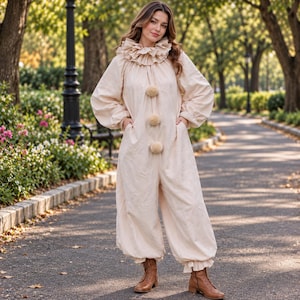 May include: A beige one-piece jumpsuit featuring a high ruffled collar, long sleeves with gathered cuffs, and three large beige pom-poms down the center front. The garment has a loose, relaxed fit with cuffed pant legs and is paired with brown boots.