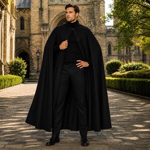 May include: A floor-length black cape worn over a black turtleneck sweater and black trousers. The cape features a single button closure at the neck, a structured collar, and a full, flowing silhouette that drapes down to the ankles.