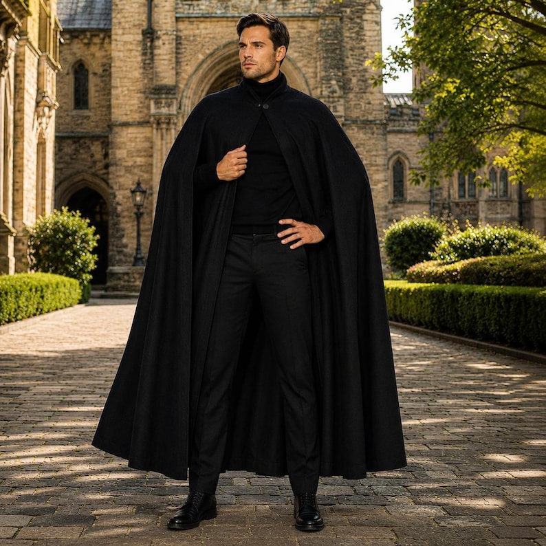 May include: A floor-length black cape worn over a black turtleneck sweater and black trousers. The cape features a single button closure at the neck, a structured collar, and a full, flowing silhouette that drapes down to the ankles.