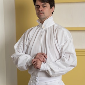 May include: White historical shirt with a high collar and buttoned cuffs. The shirt has a gathered design at the shoulders and sleeves. The matching white pants have a high waist and button details. The outfit is reminiscent of historical attire.