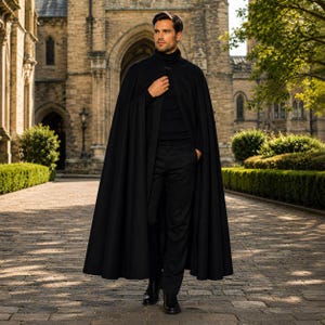 May include: A black floor-length cape worn over a black turtleneck sweater and black trousers. The cape features a high collar with a single button closure at the neck, creating a structured silhouette with long, flowing fabric panels.