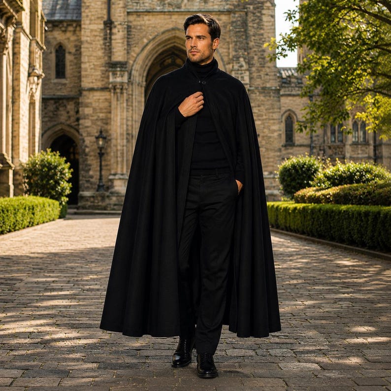 May include: A black floor-length cape worn over a black turtleneck sweater and black trousers. The cape features a high collar with a single button closure at the neck, creating a structured silhouette with long, flowing fabric panels.