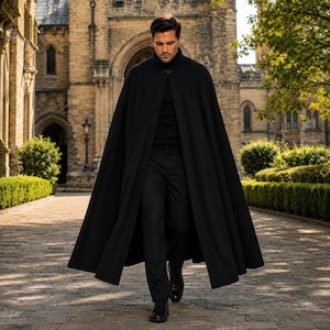 May include: A floor-length black cape featuring a high collar and a single button closure at the neck. The garment is worn over a black turtleneck sweater and black trousers, creating a monochromatic look with a structured, flowing silhouette.