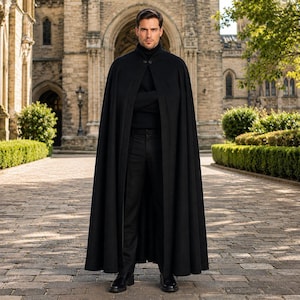 May include: A floor-length black cape featuring a high collar with two visible buttons at the neck. The cape is worn over a black turtleneck sweater and black trousers, creating a solid dark ensemble with a structured, flowing silhouette.