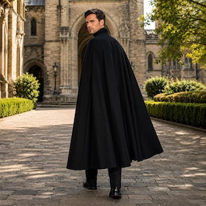 May include: A floor-length black cape with a high collar, worn over dark trousers and black dress shoes. The garment features a structured shoulder design and a full, flowing silhouette that drapes down to the ankles, creating a clean, solid appearance.