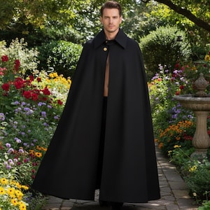 May include: A long, black cape with a wide collar and a single gold button closure. The cape is worn outdoors in a garden setting with colorful flowers and a stone fountain in the background. The cape is made of a smooth, flowing fabric.