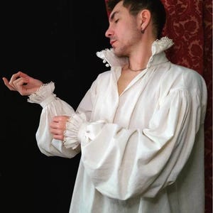 May include: A long-sleeved white shirt featuring a high standing collar with lace trim, voluminous bishop sleeves, and matching lace-trimmed cuffs. The shirt has a button-down front closure and is constructed from a solid, light-colored fabric.