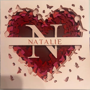May include: A layered paper art piece featuring a heart shape filled with red and pink butterflies. The letter "N" and the name "Natalie" are cut out in white. Additional butterflies are scattered around the heart.