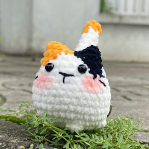 May include: A handmade crochet cat with a white body, black and orange markings, and pink cheeks. The cat has black button eyes and a stitched mouth. The cat is sitting on a bed of green plants.