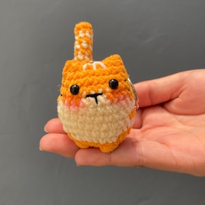 May include: A handmade orange and white crocheted cat with black eyes and a small tail. The cat has a cream-colored muzzle and pink cheeks. The cat is held in a person's hand.