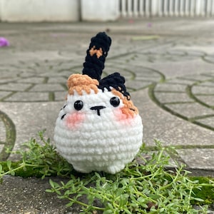 May include: A handmade crochet cat toy. The cat has a white body, black and orange ears, and a black tail. It has black eyes and pink cheeks. The toy is sitting on a stone surface with green plants.