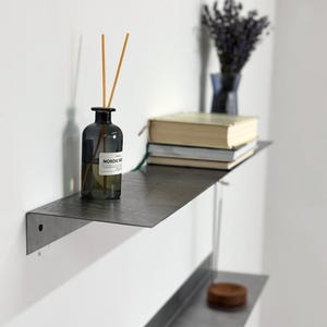 May include: Two minimalist metal shelves mounted on a white wall. The top shelf holds a dark glass diffuser bottle with reeds and a stack of books. The bottom shelf has a small wooden disc. A vase of purple flowers sits on the top shelf.