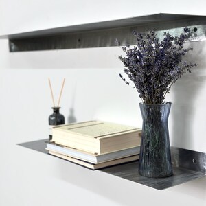 May include: Two horizontal metal wall shelves with a brushed finish. The lower shelf holds a stack of three books, a small dark glass bottle with reed diffuser sticks, and a ribbed blue glass vase filled with a bouquet of dried purple lavender.